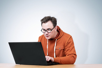 Man  in a sweatshirt and round glasses sits at a table and works with a laptop