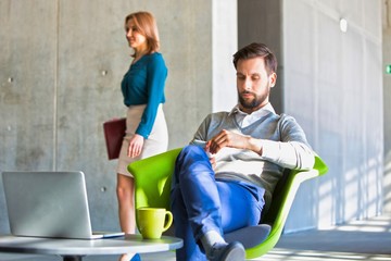 Young businessman using smartphone against mature businesswoman walking in office