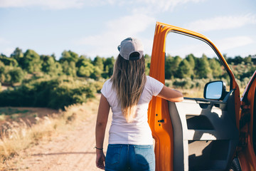 Attractive blonde girl from behind holding orange car door outdoors with mountains background