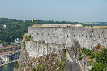 Fototapeta na wymiar Dinant, Belgium - June 26, 2019: Southern gray stone ramparts of Citadelle above the city with Belgian flag under blue sky. Green forests in back. Meuse River down.