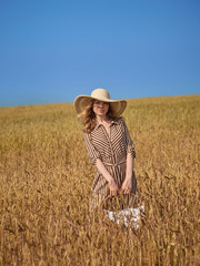 A blonde in a wheat field walks with a wicker basket of apples in a lime hat and a vertical striped dress.