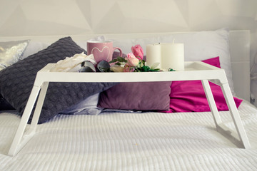 A white breakfast table on white bed with cup, flowers and candles on it. Nearby are table with flowers, in cold colors