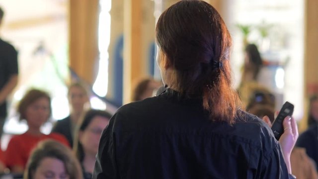 Speaker is giving speech at business event. Adult businesswoman in modern conference hall. Business and entrepreneurship seminar. Back view.