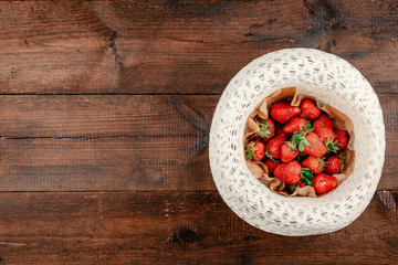 Fresh organic strawberries on round straw hat on brown rustic wooden background. Red berries on table flat lay