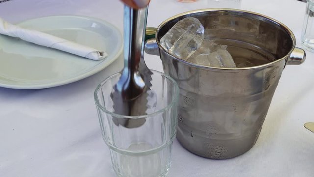 Serving ouzo on a small glass, diluted with ice cubes from an ice bucket. Pouring strong alcoholic drink ouzo on the rocks, served on a Greek tavern table with white linen tablecloth.