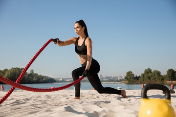 Young healthy woman doing exercise with the ropes at the beach. Single caucasian female model practicing at the river side in sunny day. Concept of healthy lifestyle, sport, fitness, bodybuilding.