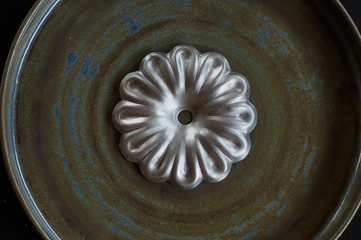 A decorative, stamped from metal flower of silver color lies on a textured, swamp color, ceramic plate close-up.