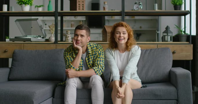 Young couple is watching TV, sitting on sofa in living room, woman looks excited, watching best TV program, man is bored. Woman tries to raise mood of man. Caucasian, white. 4K, shot on RED camera.