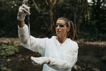 Female scientist biologist and researcher in protective suit taking water samples from polluted river.
