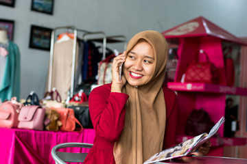 portrait of a young muslim entrepeneur sitting and smile talking in her smartphone in front of fashion boutique as background -image