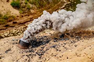 Fototapeta premium Grey smoke comes out from smoke bomb. The bomb on sand near a forest. Military training. Air pollution