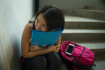 Dramatic portrait of Asian female college student bullied. Young depressed and sad Chinese girl sitting lonely on campus staircase suffering bullying and harassment