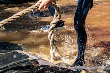 A man overcomes an obstacle at a sport event. He comes up from water and holds rope. Sport activity in the forest