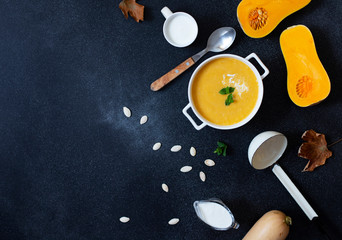 Autumn Pumpkin Cream soup on a black background, served with cream, pumpkin seeds and mint. Cut pumpkin, autumn leaves, soup ladle, white plate. Top view. Copyspace