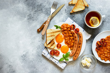 English breakfast with fried sausages, beans, mushrooms, fried eggs, grilled cherry tomatoes. Served with a cup of tea with lemon, bread toast and butter. Gray background, top view, copyspace