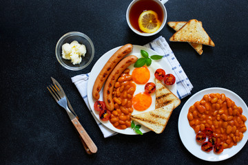 English breakfast with fried sausages, beans, mushrooms, fried eggs, grilled cherry tomatoes. Served with a cup of tea with lemon, bread toast and butter. Black background, top view, copyspace
