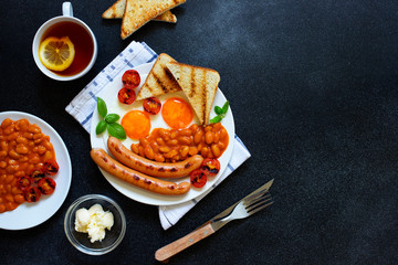 English breakfast with fried sausages, beans, mushrooms, fried eggs, grilled cherry tomatoes. Served with a cup of tea with lemon, bread toast and butter. Black background, top view, copyspace