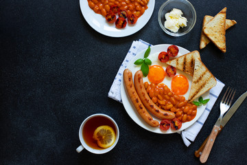 English breakfast with fried sausages, beans, mushrooms, fried eggs, grilled cherry tomatoes. Served with a cup of tea with lemon, bread toast and butter. Black background, top view, copyspace