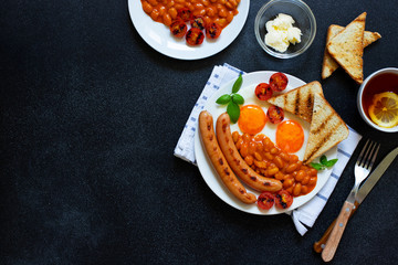 English breakfast with fried sausages, beans, mushrooms, fried eggs, grilled cherry tomatoes. Served with a cup of tea with lemon, bread toast and butter. Black background, top view, copyspace