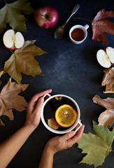 Obraz na płótnie Canvas Female hands holding mug with mulled wine. Autumn and winter warm alcoholic drink. Autumn leaves, honey, apples, oranges, vintage dishes. Autumn cozy mood. Flat lay, dark background. Vertical