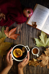 Female hands holding a mug of hot mulled wine. Mulled wine, honey, autumn leaves, apples, a book and a woolen sweater on a wooden background. Autumn cozy mood. Autumn background. Flat lay. Vertical