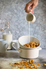 Obraz na płótnie Canvas Granola with nuts and raisins in a white bowl. Healthy breakfast served on a light table. Milk is pouring into a bowl from a cup. Female hand is pouring milk. Light neutral tones. Vertical