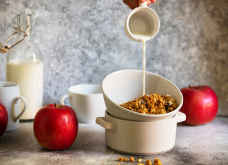 Granola with nuts and raisins in a white bowl. Healthy breakfast served on a light table with red apples. Milk is pouring into a bowl from a cup. Female hand is pouring milk.