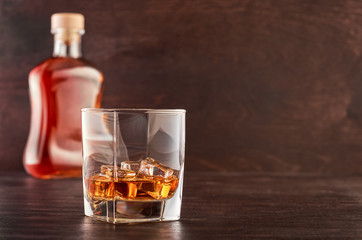A glass of whisky with ice on a wooden table, a full bottle of whisky in the background