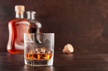 A glass of whisky with ice on a wooden table, two bottles of whisky in the background, full and open