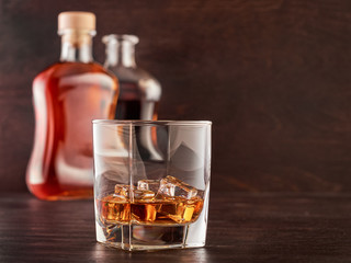 A glass of whisky with ice on a wooden table, two bottles of whisky in the background, full and open