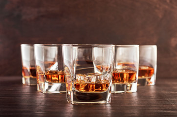 A set of six glasses of whiskey with ice standing on a wooden table. One glass in the foreground, the other glasses in the background with different depth of field.