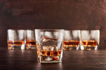 A set of six glasses of whiskey with ice standing on a wooden table. One glass in the foreground, the other glasses in the background with different depth of field.