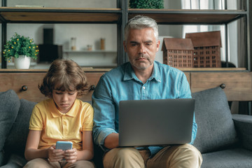 Father using laptop, sitting near son with smartphone
