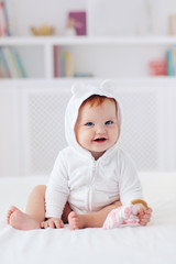 happy infant baby girl in funny ear hood is sitting on the bed at home