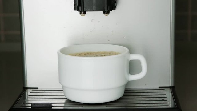 delicious coffee drops fall from machine dispenser into white cup with brown foam on support zoom in extreme close view