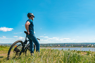 A male cyclist stands on a hill and looks at the city. A man in a helmet and with a bicycle on nature with his back to the camera.