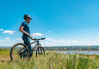 Fototapeta premium A male cyclist stands on a hill and looks at the city. A man in a helmet and with a bicycle on nature with his back to the camera.