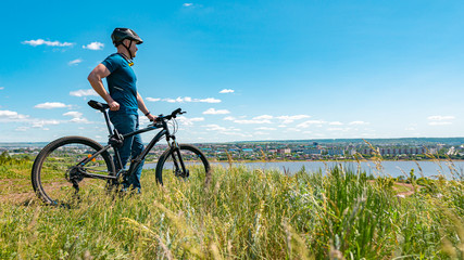 A male cyclist stands on a hill and looks at the city. A man in a helmet and with a bicycle on nature with his back to the camera.