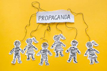 Word Propaganda, paper men and women manipulated with threads on the yellow background.