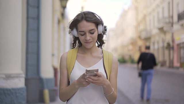 Beautiful young woman walking on the city street and listening to music, moving with it, enjoyment.