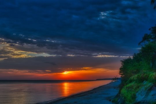 wsch&oacute;d słońca nad Bałtykiem w okolicach Dziwnowa, chmury i słońce odbijają się w spokojnej wodzie morza
