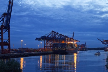 Illuminated Container Ship / Freighter at Dusk in the port of Hamburg with big Gantry Cranes (Container Cranes). Harbor Tour Boat next to it