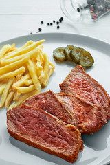 Gourmet medium rare beefsteak with French fries, pickled cucumber and grilled cherry tomatoes on a white plate with a glass of sparkling water.