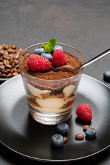 Classic tiramisu dessert portion with blueberries and raspberries in a glass cup on stone serving board on concrete background
