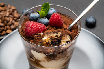 Classic tiramisu dessert portion with blueberries and raspberries in a glass cup on stone serving board on concrete background