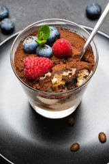 Classic tiramisu dessert portion with blueberries and raspberries in a glass cup on stone serving board on concrete background