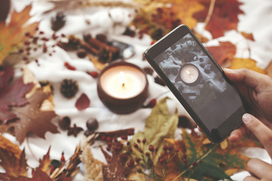 Autumn hygge flat lay. Hand holding phone and taking photo of candle, berries, fall leaves, anise,herbs, acorns, nuts, cinnamon, cotton on white textile. Hygge lifestyle, cozy autumn mood