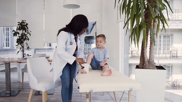 Young female doctor bandaging leg of little patient.