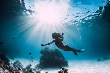 Woman freediver with fins swim over sandy sea with fish and sun rays underwater