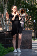Girl learning to play trombone. Girl plays standing on the alley of a city park.